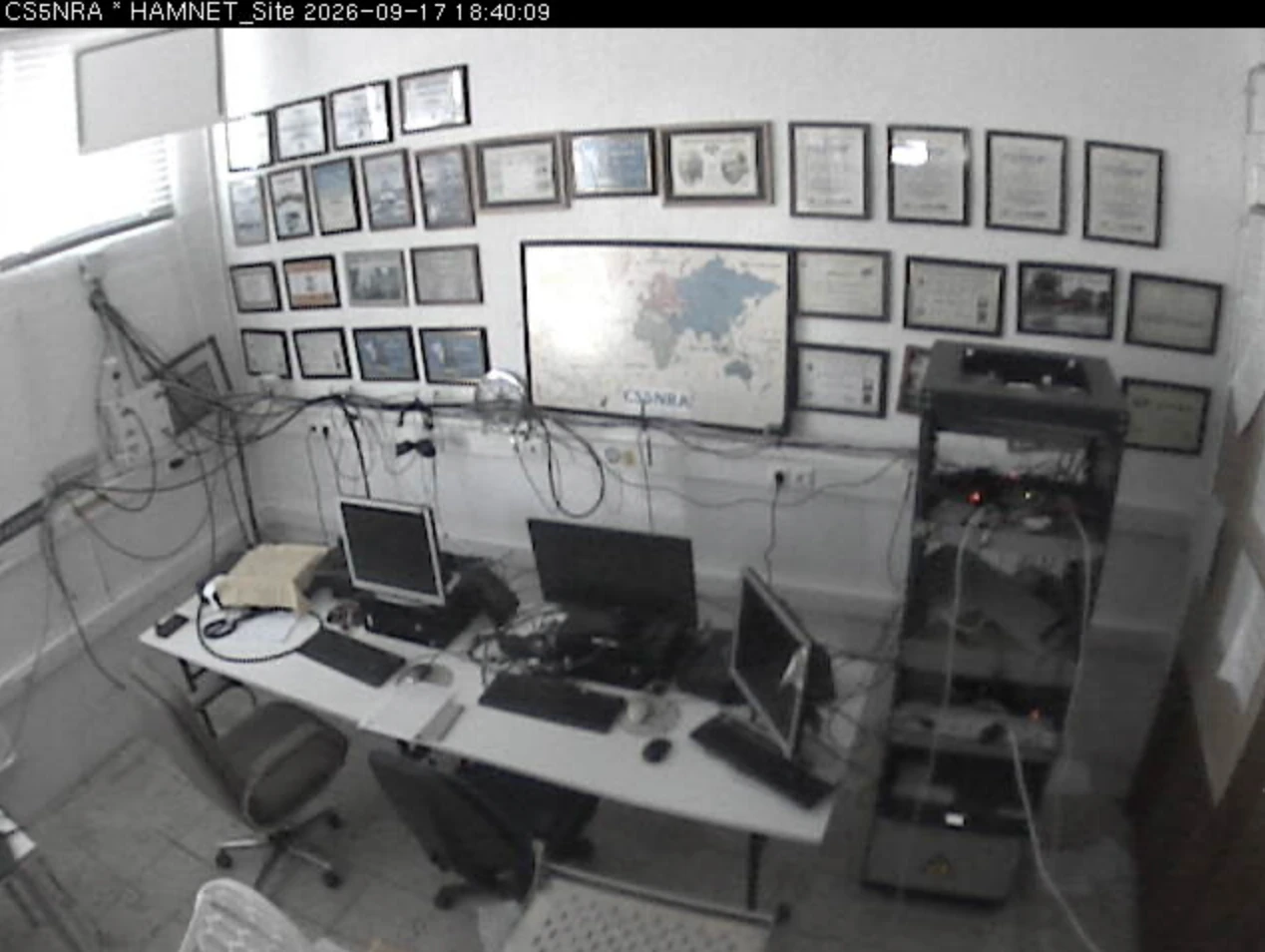 Imagem da câmara CS5NRA: sala da estação com computadores, equipamento de rádio, um bastidor e diplomas nas paredes.